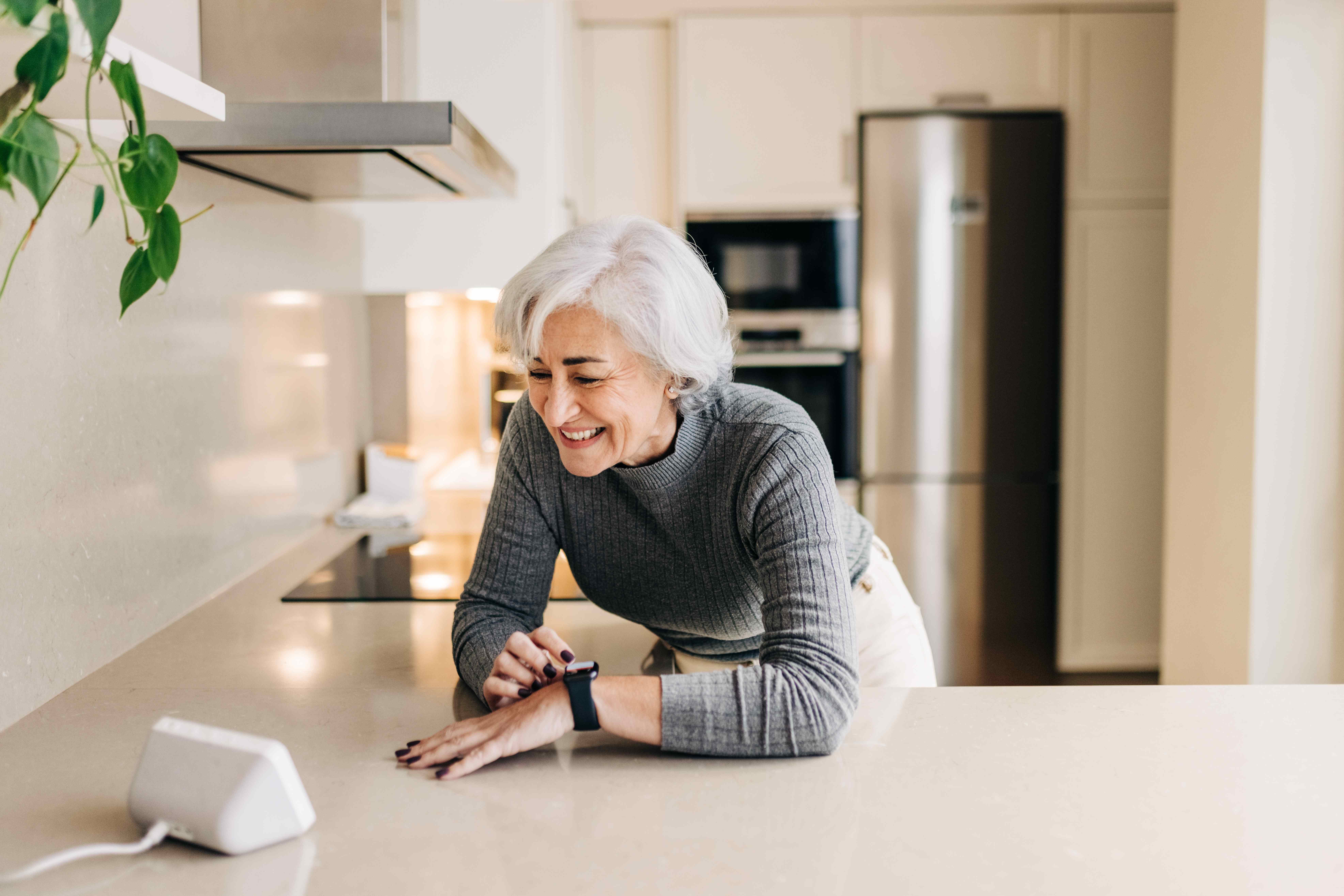 An older woman smiling while interacting with her smartwatch and a smart home hub device on her kitchen counter.